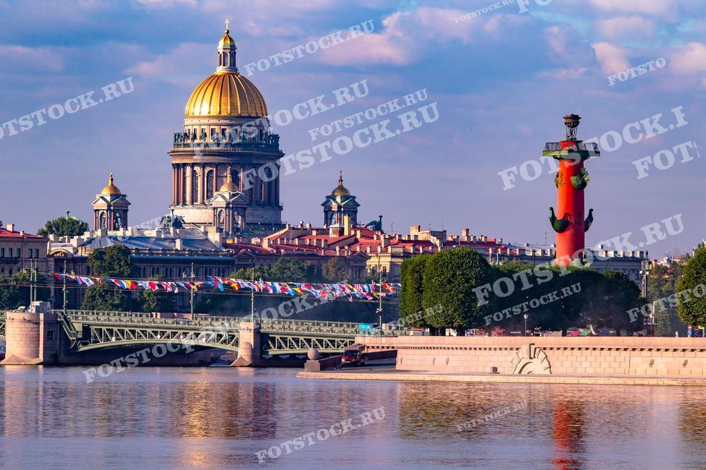 На фотографии запечатлены Исаакиевский собор и Ростральные колонны в Санкт-Петербурге. На переднем плане вода реки Невы мягко отражает архитектурные детали, а украшенный мост добавляет праздничности этому городскому пейзажу. Такой вид отлично подойдёт для использования в публикациях о Санкт-Петербурге, его культурном наследии и архитектуре, а также в качестве визуального материала для туристических проспектов и иллюстраций к статьям.